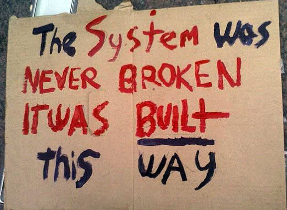 Cardboard sign saying 'The System was never broken It was Built this way
